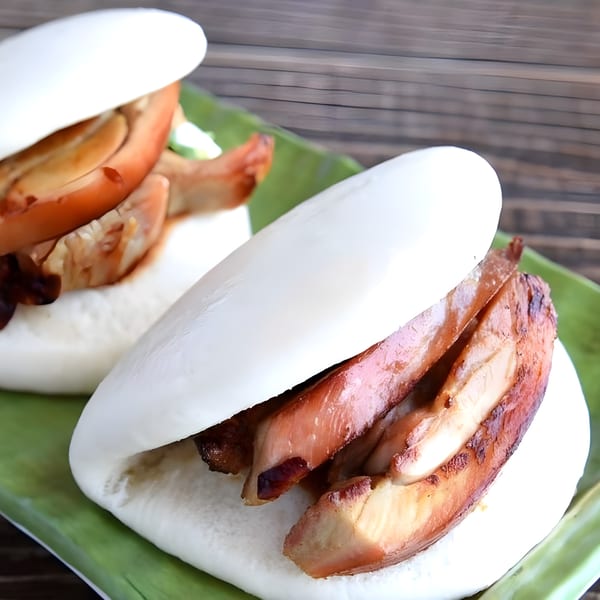Two Corner Ramen & Poke pork belly bao buns on a green leaf, against a wooden table.